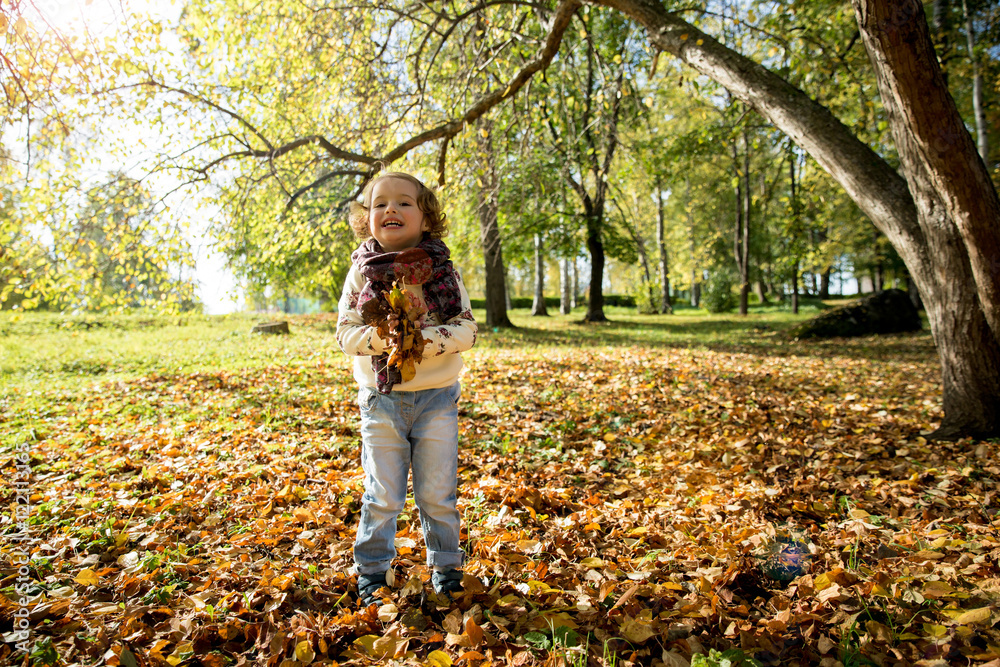 Happy cheerful kid playing in the autumn park. Little girl throwing fallen yellow leaves and