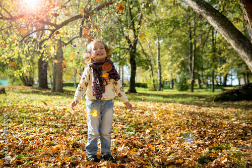 Happy cheerful kid playing in the autumn park. Little girl throwing fallen yellow leaves and laughing. Sunny weather, fresh air. Carefree childhood concept. Healthy lifestyle. © Suzi Media 