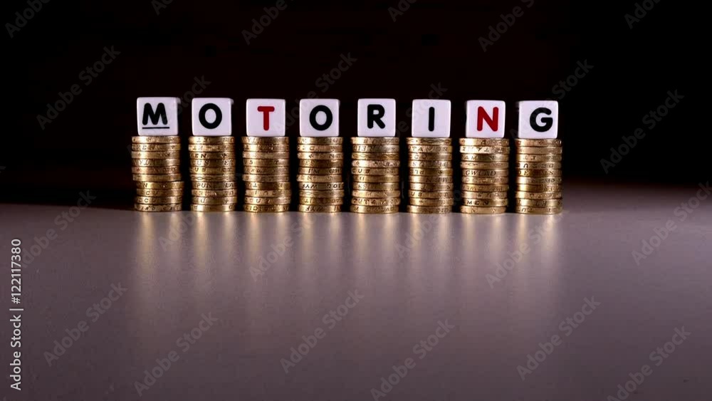 £1 sterling coin stacks with ‘motoring’ in letters on top. A model car drives past. Cost of motoring concept. £1 sterling coin stacks with ‘motoring’ in letters on top. A model car drives past. Cost of motoring concept.