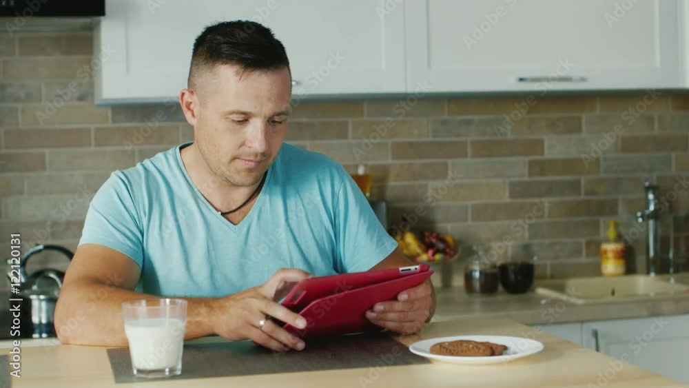 A young man uses tablet in the kitchen, standing next to a glass of milk. Healthy lifestyle A young man uses tablet in the kitchen, standing next to a glass of milk. Healthy lifestyle