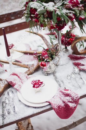 Wedding table setting in marsala colors with plates  cutlery  red floral compositions  candles  velvet napkins  deer horns on table covered with snow
