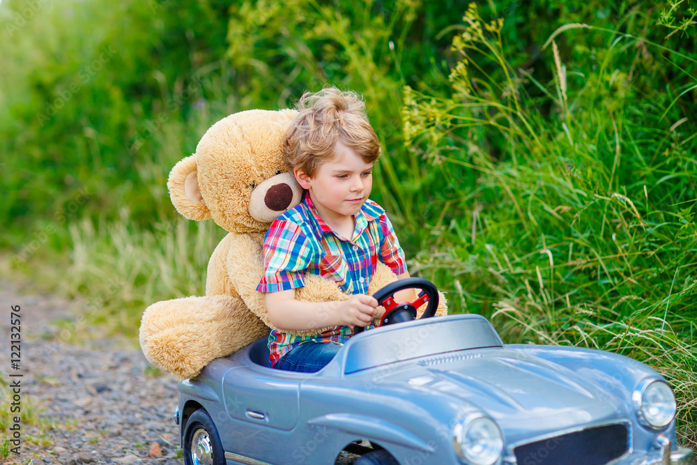 Little kid boy driving big toy car with a bear, outdoors.