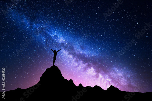Landscape with Milky Way. Colorful night sky with stars and silhouette of a standing happy woman with raised-up arms on the mountain peak on the background of beautiful galaxy. Blue milky way rocks