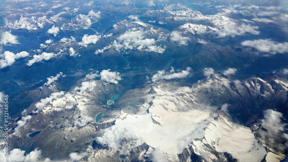 Traveling by air through an airplane window. Aerial view of Alps mountains surrounded by cirrus and cumulus clouds with little turbulence, showing Earth's atmosphere.