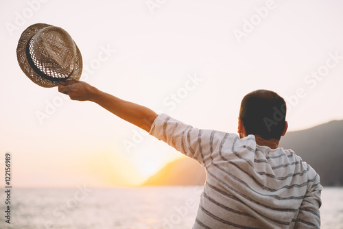 Greece, Cylcades Islands, Amorgos, man holding straw hat enjoying the sunset next to the sea photo