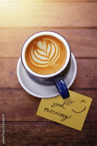 Cup of Coffee Latte on Wooden table with Paper Note and word: good morning and smilely face in Morning Sunshine, selective focus on a cup