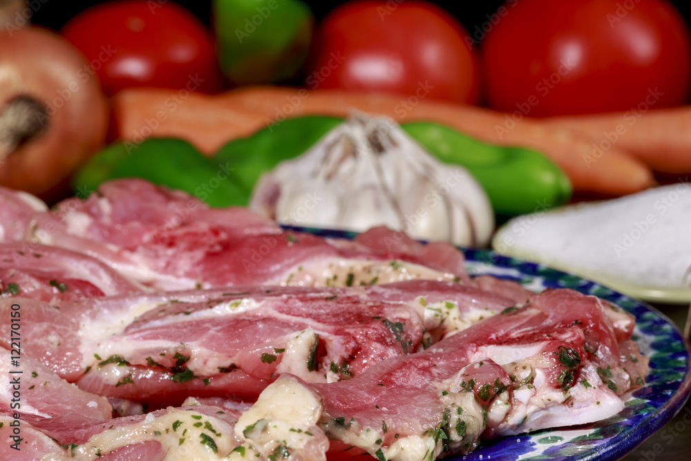 Close-up of turkey raw meat dish marinated with garlic and parsley in front of a dish with salt, garlic, onions,green peppers, tomatoes and carrots