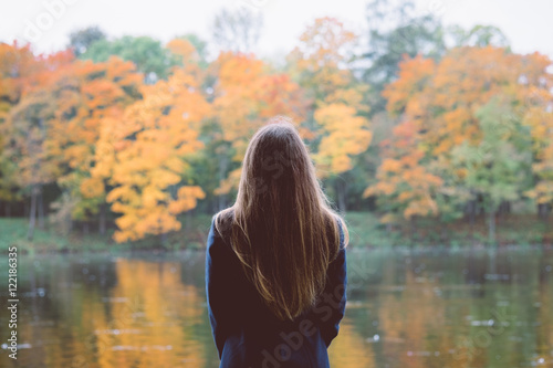 Outdoor autumn portrait of girl standing backwards with straight hair. Back view of woman explores calm lake and forest nature at fall. Image toned with old film style filters