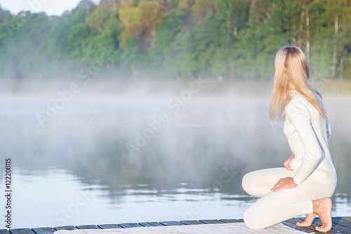 After practicing yoga  woman staring at the lake and the trees which was hidden behind a thick fog. Above the lake is a mist. White clothes.