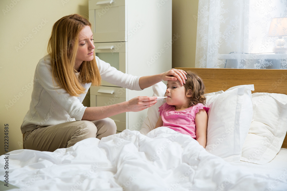Mother measuring temperature of her ill kid. Sick child with high fever laying in bed and mother holding thermometer. Hand on forehead. Mother measuring temperature of her ill kid. Sick child with high fever laying in bed and mother holding thermometer. Hand on forehead.