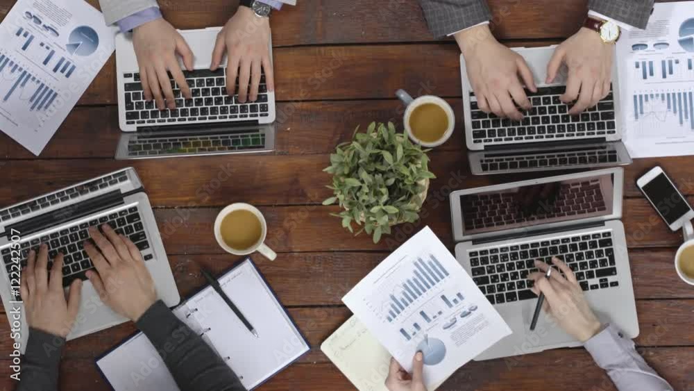 Time lapse view from the top of hands of business colleagues sitting at the meeting table, typing on laptops, working with documents and drinking coffee