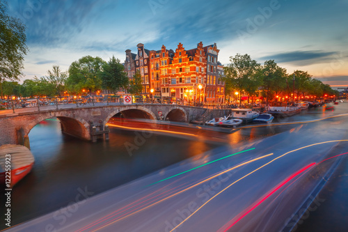 Photography Amsterdam canal, bridge and typical houses, boats and luminous track from the boat during morning twilight blue hour, Holland, Netherlands