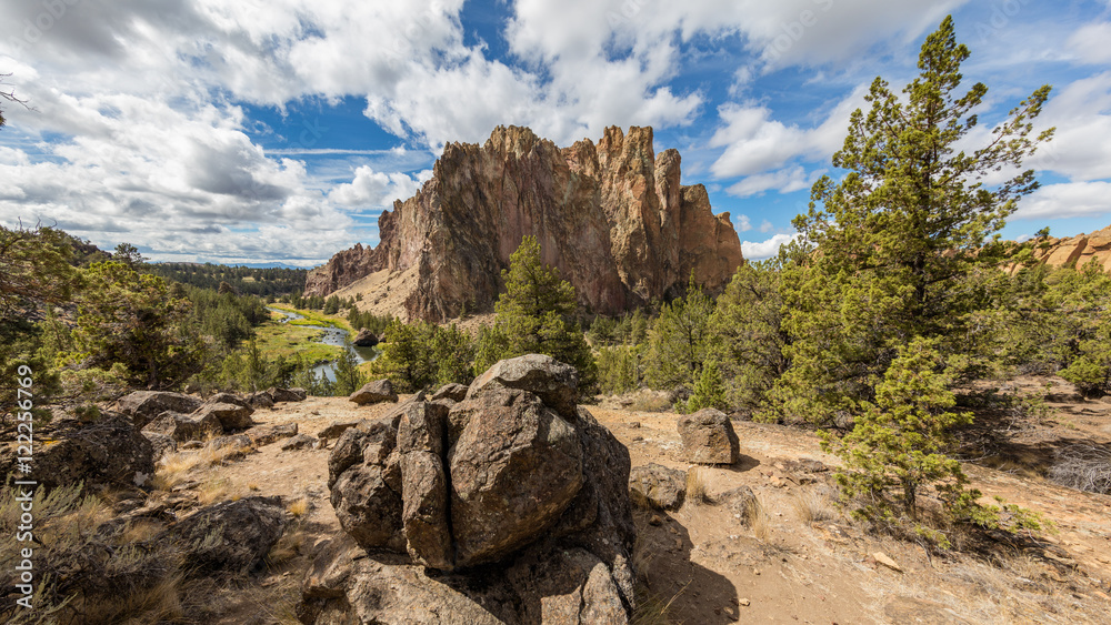 Large stones in the pines. The river flows between rocks. Beautiful landscape of yellow sharp cliffs. Dry yellow grass grows at the foot of cliffs. Smith Rock state park, Oregon