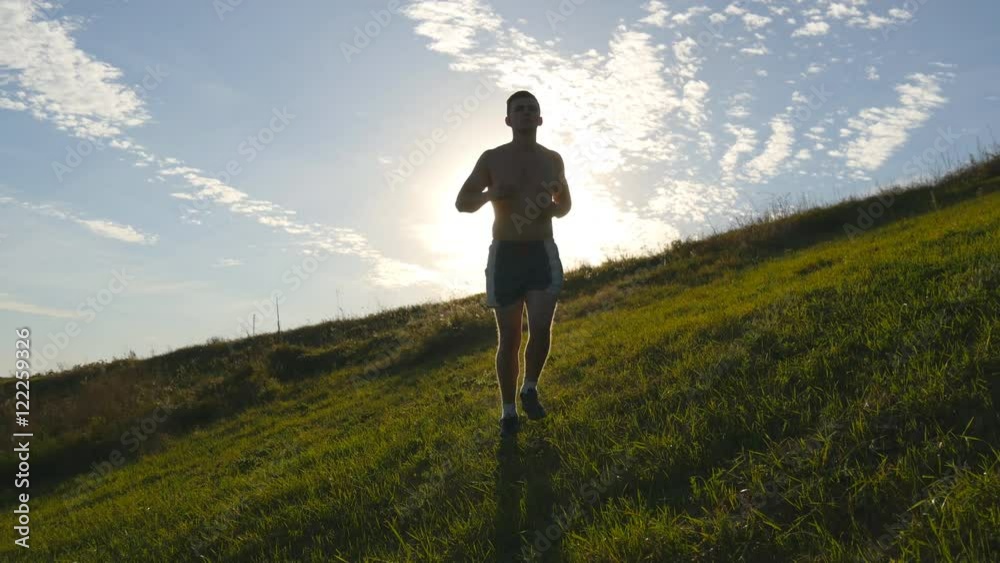 Young man running over green hill over blue sky background. Male athlete is jogging in nature at sunset. Sports runner jogging uphill outdoor at sunrise with flare. Cross-country training. Lifestyle Young man running over green hill over blue sky background. Male athlete is jogging in nature at sunset. Sports runner jogging uphill outdoor at sunrise with flare. Cross-country training. Lifestyle