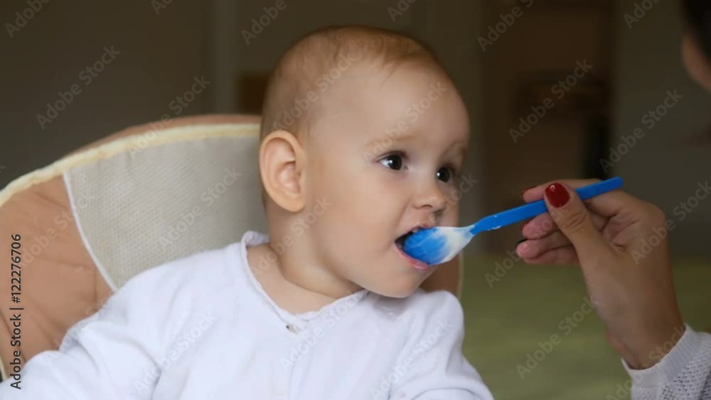 Little girl eating yogurt in Child seat and smiling. Happy family concept. Mother with spoon and baby food. Little girl eating yogurt in Child seat and smiling. Happy family concept. Mother with spoon and baby food.