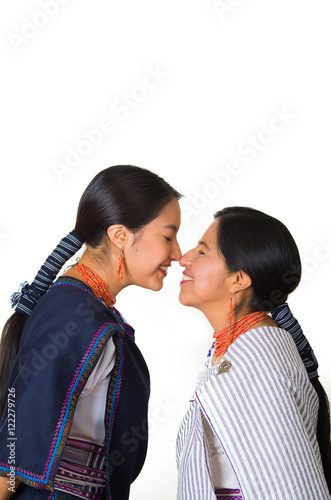Beautiful hispanic mother and daughter wearing traditional andean clothing, seen from profile angle facing each other touching noses smiling, white studio background