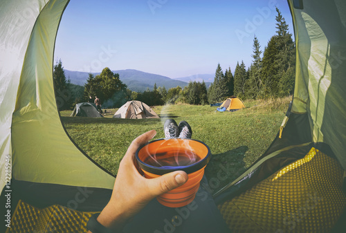 Woman lying in a tent with coffee ,view of camping with  people near the fire   and forest © natalia_maroz