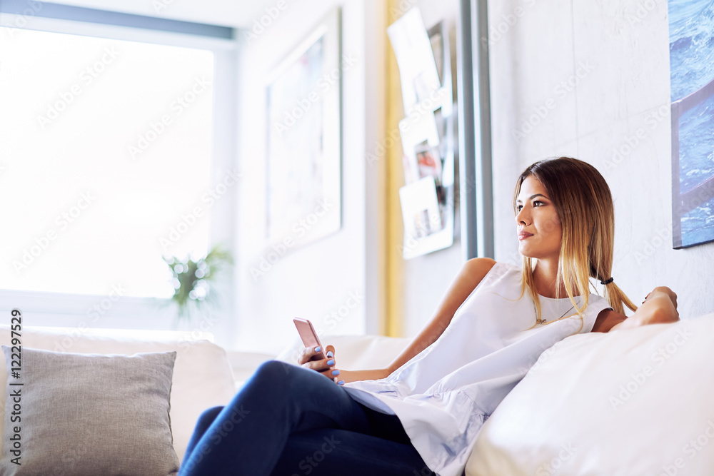 young girl sitting on a sofa and looking thoughtfully right while holding the phone in your hand young girl sitting on a sofa and looking thoughtfully right while holding the phone in your hand