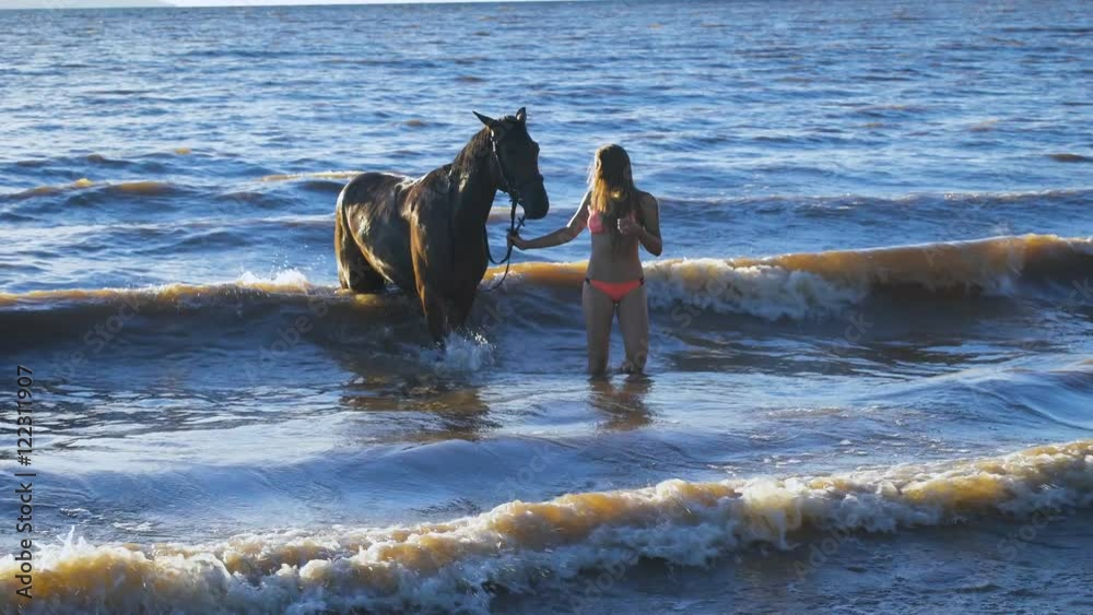 A beautiful blonde young sexy woman walking next to a horse at a lake A beautiful blonde young sexy woman walking next to a horse at a lake
