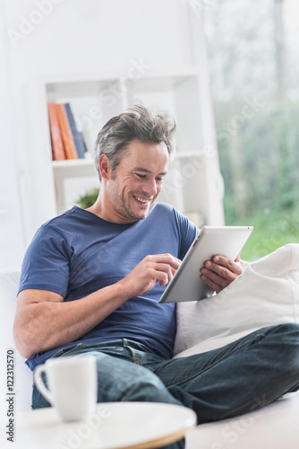  cheerful man at home using a tablet, he sits on a white couch