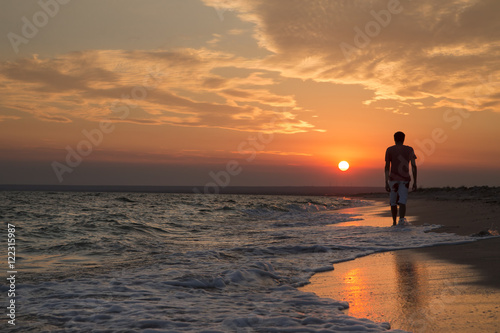 silhouette of a young man who walks on the beach, against the backdrop of a beautiful sunset © YURII Seleznov