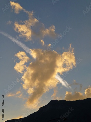 Clouds and vapour trail illuminated by setting sun with mountain peak silhouetted against the sky