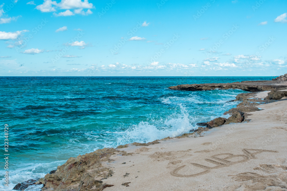 Beautiful view of rocky coast of azure coastline of Caribbean Sea in hot sunny summer day. Quiet small waves are splashing against coastal stones. Inscription Cuba on the sand. Clear serene blue sky Beautiful view of rocky coast of azure coastline of Caribbean Sea in hot sunny summer day. Quiet small waves are splashing against coastal stones. Inscription Cuba on the sand. Clear serene blue sky