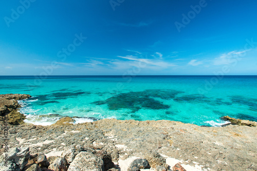 Beautiful view of the rocky coast of the azure coastline of the Caribbean Sea in hot sunny summer day. Quiet small waves are splashing against coastal stones. Clear serene blue sky