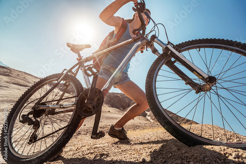 Young lady climbing up the sandy hill with a bicycle in the desert at sunny day