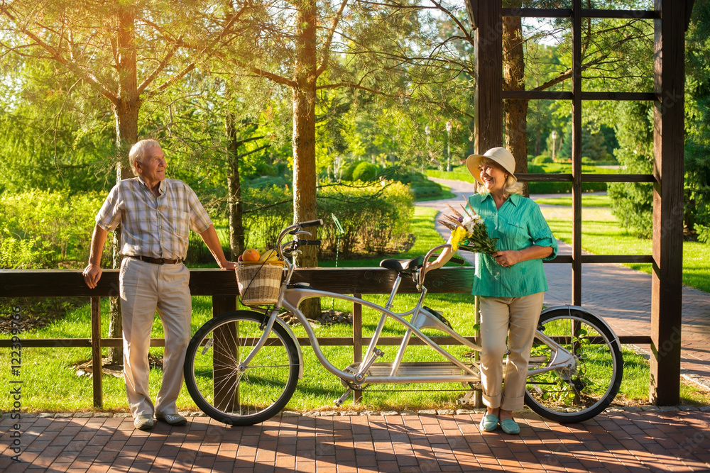 Senior couple near tandem bike. Woman smiling and holding bouquet. How about a picnic. Surprise for wife. Senior couple near tandem bike. Woman smiling and holding bouquet. How about a picnic. Surprise for wife.