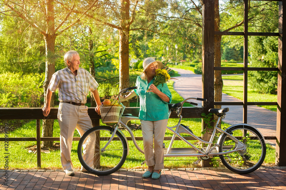 Obraz premium Senior couple and tandem bicycle. Woman is smelling bouquet. Her favorite flowers. Small surprise for closest person.
