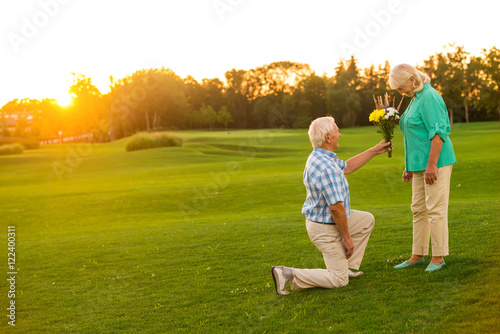Man with bouquet is kneeling. Couple of seniors on meadow. This is for you, darling. Ho to impress a woman.