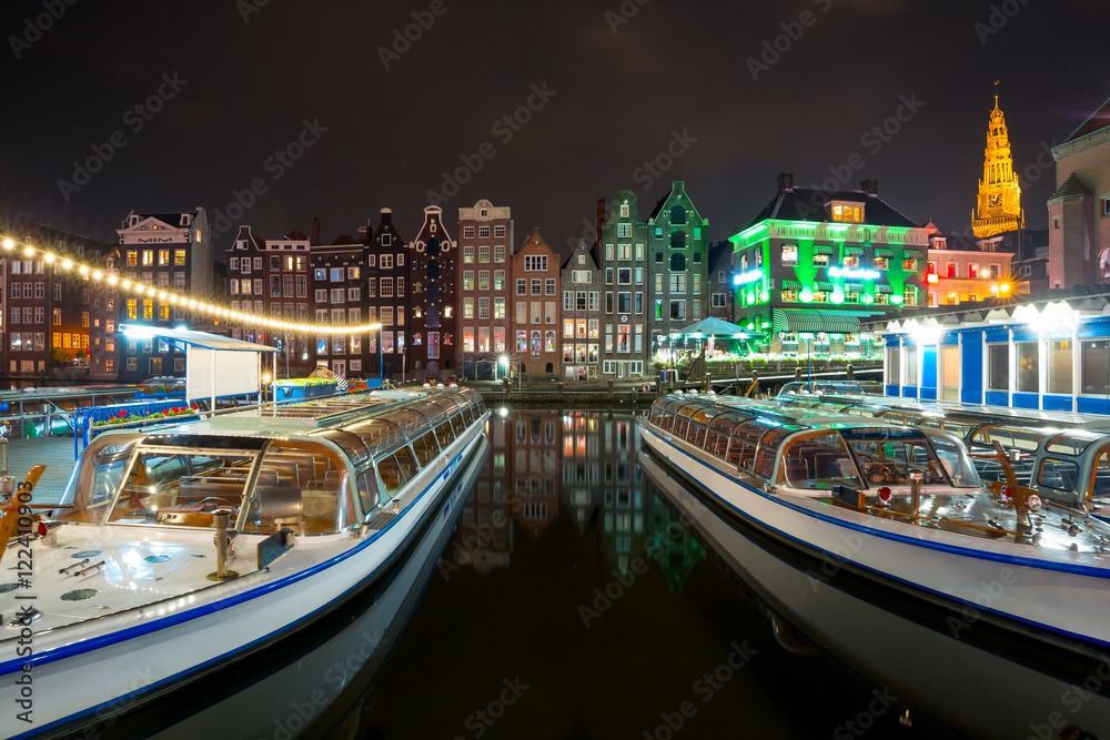 Beautiful typical Dutch dancing houses, Oude Kerk church and tourist boats at the Amsterdam canal Damrak at night, Holland, Netherlands.