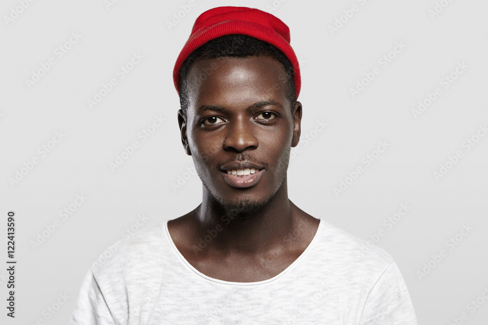 Open and sociable. Handsome happy young Afro-American hipster wearing red trendy hat, smiling, showing his white teeth. Black student dressed casually relaxing indoors, looking joyful and cheerful Open and sociable. Handsome happy young Afro-American hipster wearing red trendy hat, smiling, showing his white teeth. Black student dressed casually relaxing indoors, looking joyful and cheerful