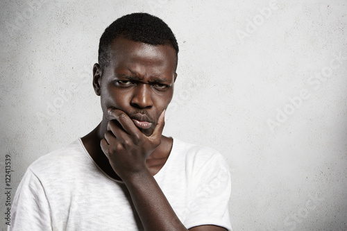 Portrait of skeptical African young man looking with suspicious or annoyed expression, holding hand on chin, doubting, thinking over something. Black male with disgust or disapproval on his face