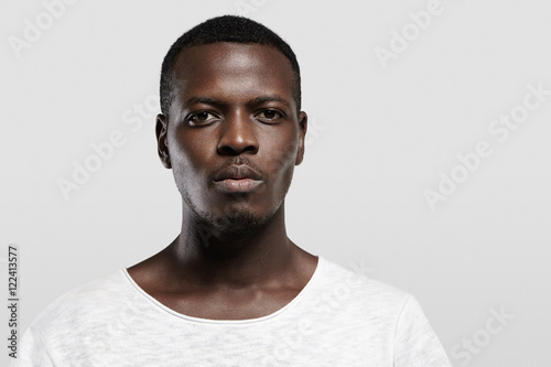 Headshot of attractive serious African student with small beard and moustache, dressed in casual t-shirt, looking at camera with confident and thougthful expression on his face standing at gray wall photo