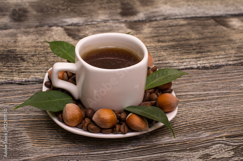 small white cup of coffee with cocoa beans, hazelnuts and green leaves on wooden background