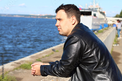 Man in leather jacket stands near river with ship, looks away an