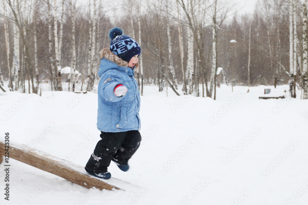 Happy little boy runs on log in park during snowfall at winter d
