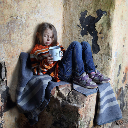 Sad little girl with dirty face sits on old blanket in basement with iron mug in hands - refugee orphan, homelessness, poverty concept