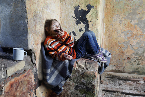 Tableau sur toile Sad little girl with dirty face sits on old blanket in basement and eats rough b
