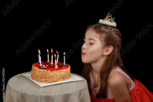 Princess anniversary - cute girl in red dress and crown on head going to blow out the candles on birthday cake - low key side view on black background