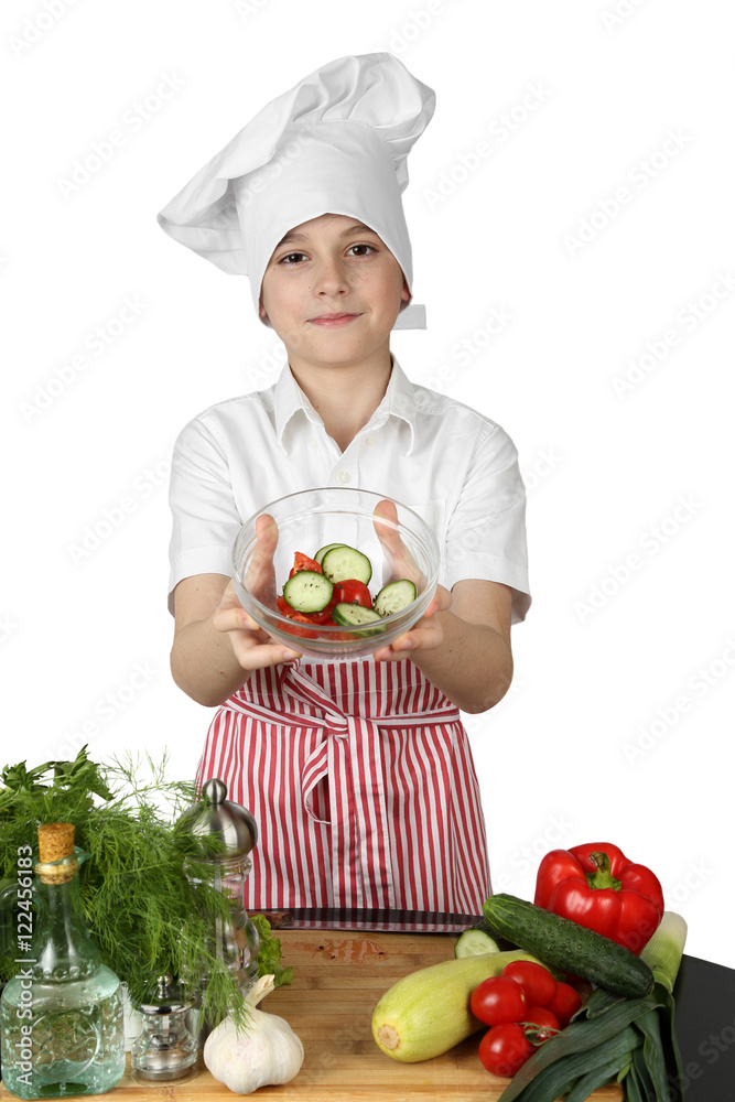 Teenage boy in cook chef hat and apron holds bowl of salad in hands above table with vegetables and condiments isolated on white background - vegetarian cuisine Teenage boy in cook chef hat and apron holds bowl of salad in hands above table with vegetables and condiments isolated on white background - vegetarian cuisine
