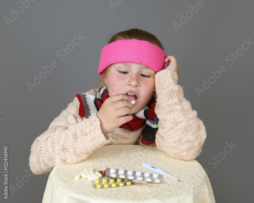 Sick in headband girl sits near table with medicine and take pill on gray background - Treatment, cold or disease concept