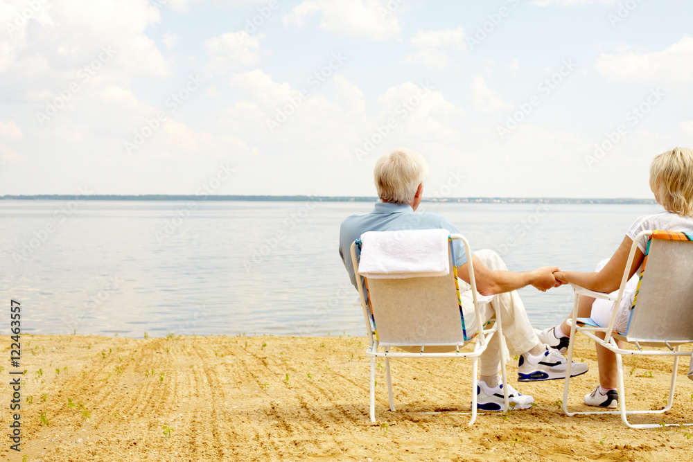 Rear view of senior couple resting in chaise lounges on lakeside and holding hands