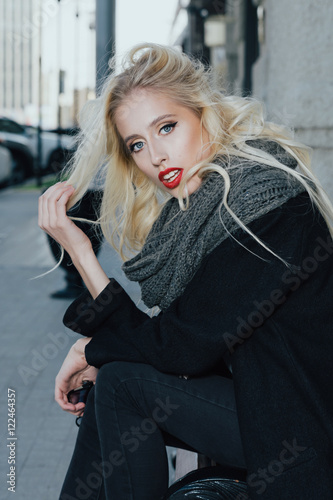Woman with blond curly hair on bench. Beautiful Portrait of sensual young model wearing a coat and grey scarf. With glamour red lips make-up, eye arrow makeup, purity skin
