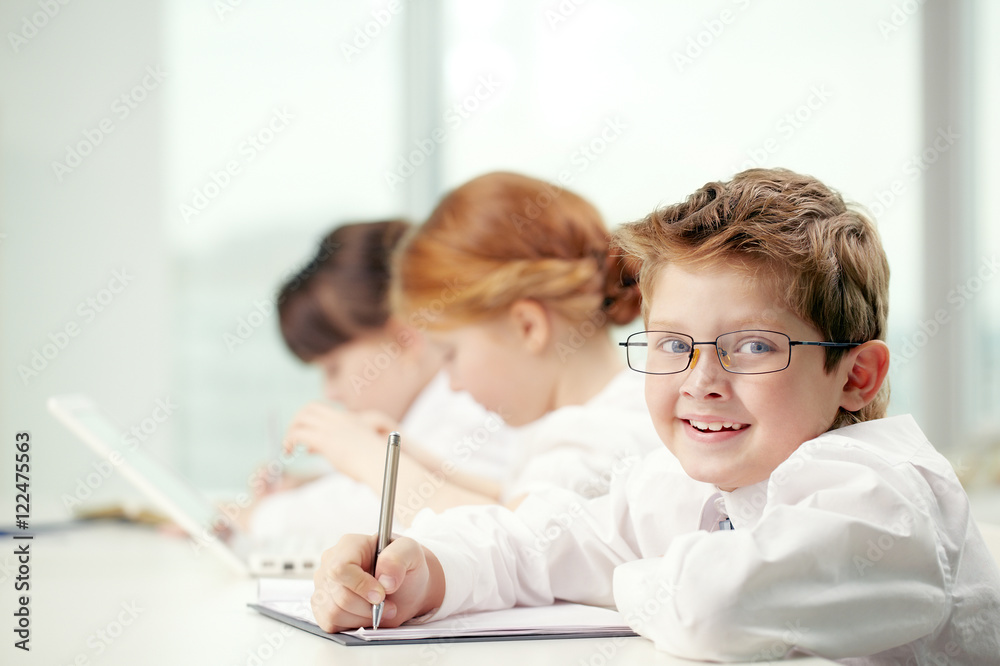Little boy in glasses looking at camera and smiling by his classmates Little boy in glasses looking at camera and smiling by his classmates