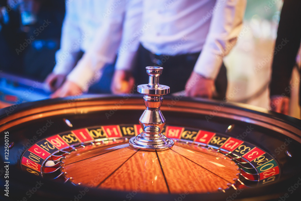 A close-up vibrant image of multicolored casino table with roulette in motion, with casino chips. the hand of croupier, mone and a group of gambling rich wealthy people in the background
A close-up vibrant image of multicolored casino table with roulette in motion, with casino chips. the hand of croupier, mone and a group of gambling rich wealthy people in the background