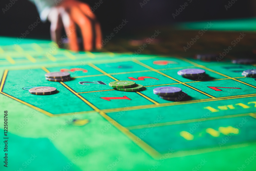 A close-up vibrant image of multicolored casino table with roulette in motion, with the hand of croupier, and a group of gambling rich wealthy people in the background
A close-up vibrant image of multicolored casino table with roulette in motion, with the hand of croupier, and a group of gambling rich wealthy people in the background