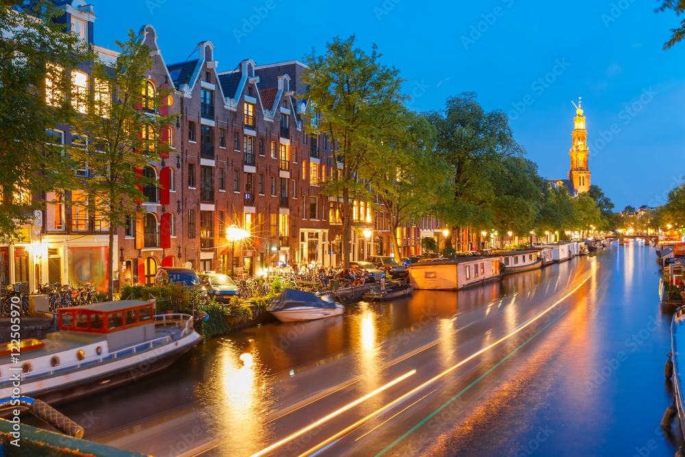 Fototapeta premium Night city view of Amsterdam canal Prinsengracht with houseboats and Westerkerk church and luminous track from the boat, Holland, Netherlands.